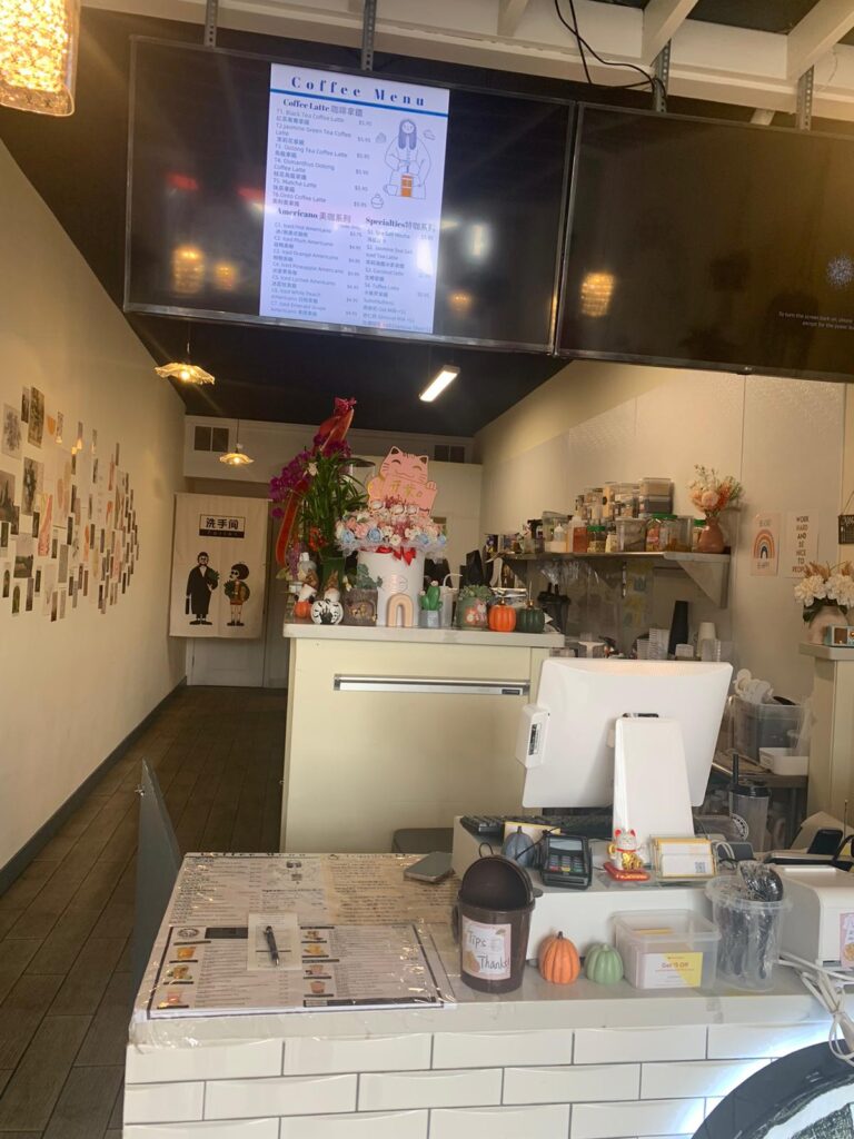 The cashier section of the coffee spot. Menus are displayed on overhead TVs or on physical paper. Adorned with the cutesy aesthetic of minimalistic white and pastel whites and pinks